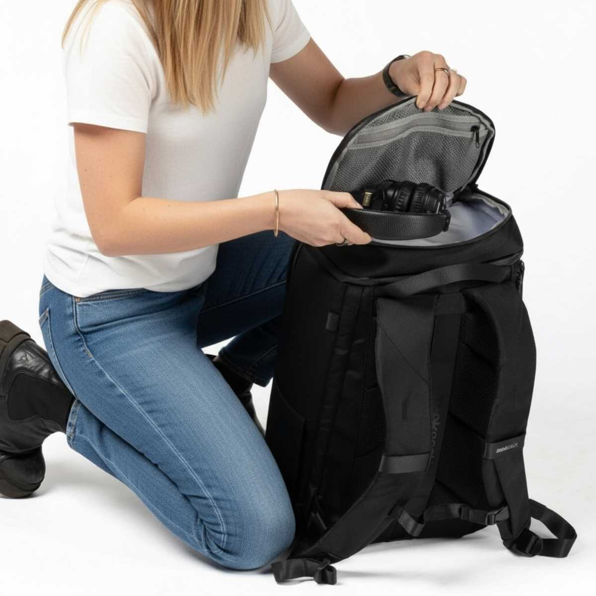 Nainen valkoisessa t-paidassa ja farkuissa polvillaan lattialla, avaa mustan repun yläosaa.