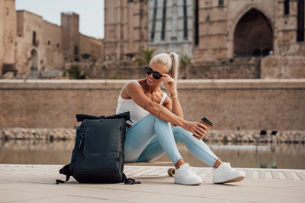 Nainen istuu ulkona Rolltop-repun kanssa, juo kahvia ja hymyilee, taustalla vanha kivirakennus.