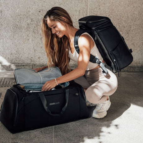 Nainen kyykyssä ja pakkaa Zeeksack-weekenderia, selässä musta reppu, valo osuu kasvoihin.