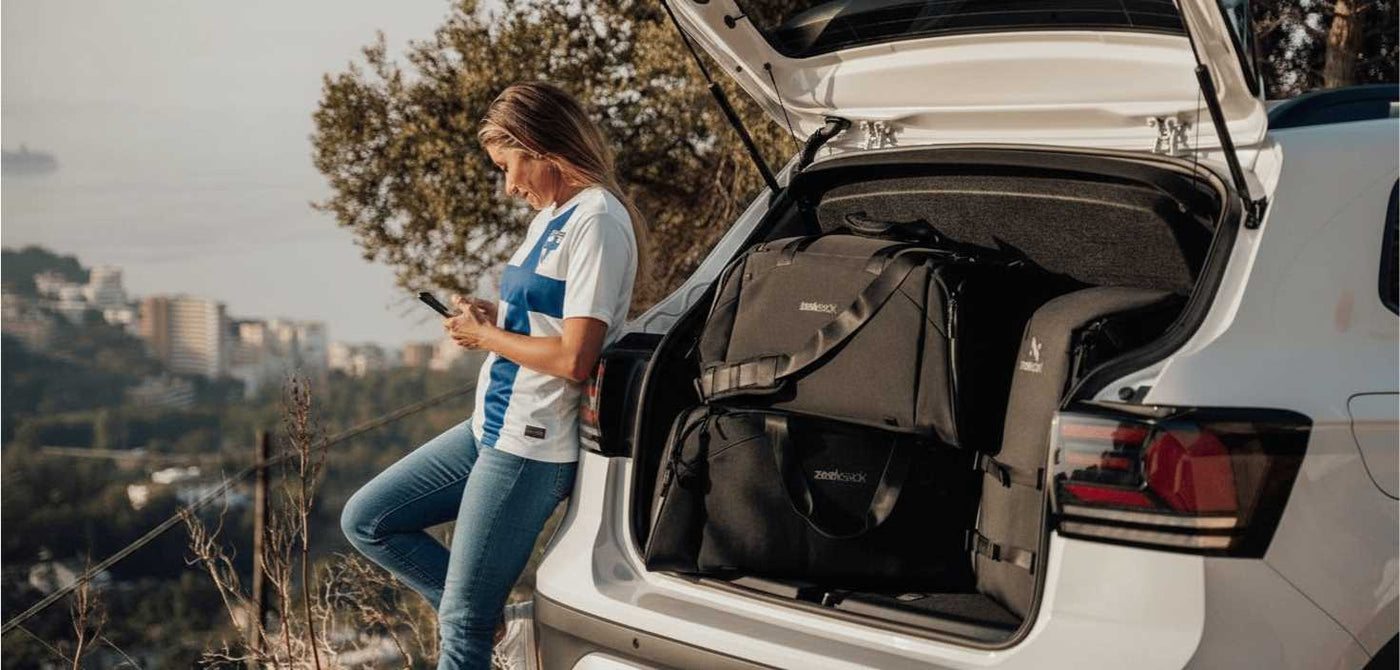 Nainen sinisissä farkuissa, valkoisissa lenkkareissa ja sinivalkoisessa Suomen maajoukkuetyylisessä paidassa nojaa autoon, jonka takaluukku on auki. Tavaratilassa on mustia Zeeksack-matkalaukkuja ja reppu. Taustalla näkyy kaupunki ja meri.